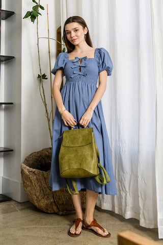 A woman in a blue, short-sleeved knee-length dress stands indoors, holding the Convertible Classic Fashion Backpack in green. She wears brown sandals on a polished floor, with a large woven basket and plants behind her.